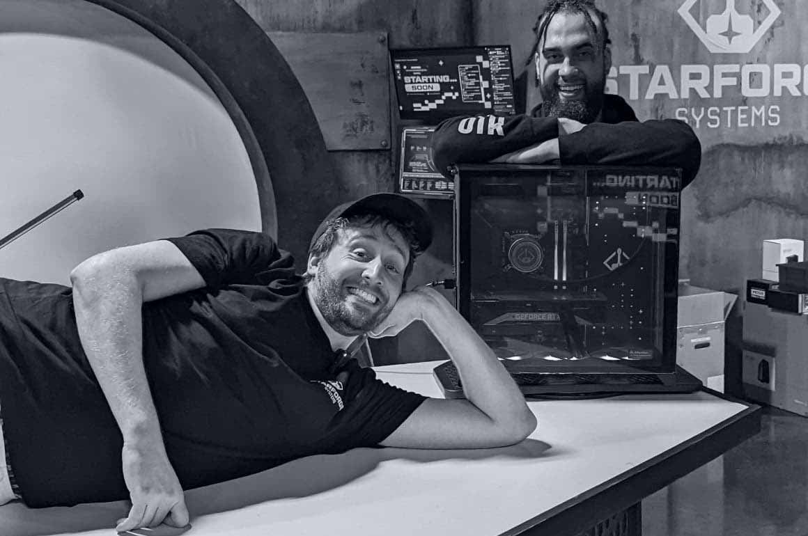 Two men pose for a photo in a studio. One on top of a table, lying on their side propped up on one elbow with the hand supporting the head and the other stands behind and leans on a Starforge PC.